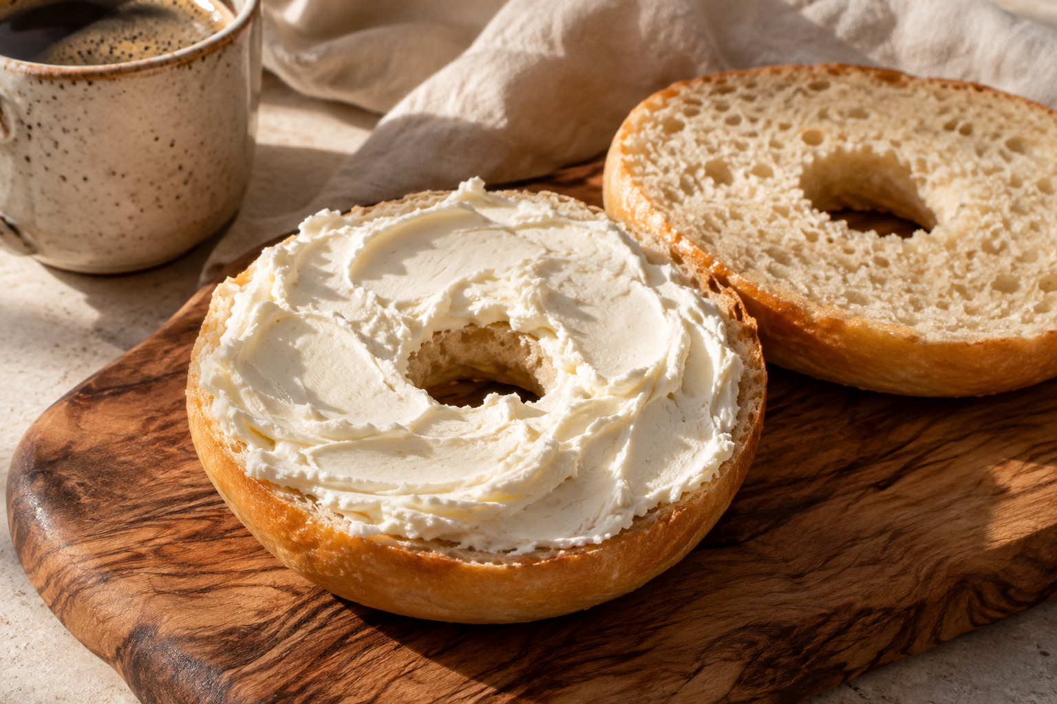 Un bagel nature tranché, garni de fromage frais aux sillons crémeux, sur une planche en noyer avec un café et du lin
